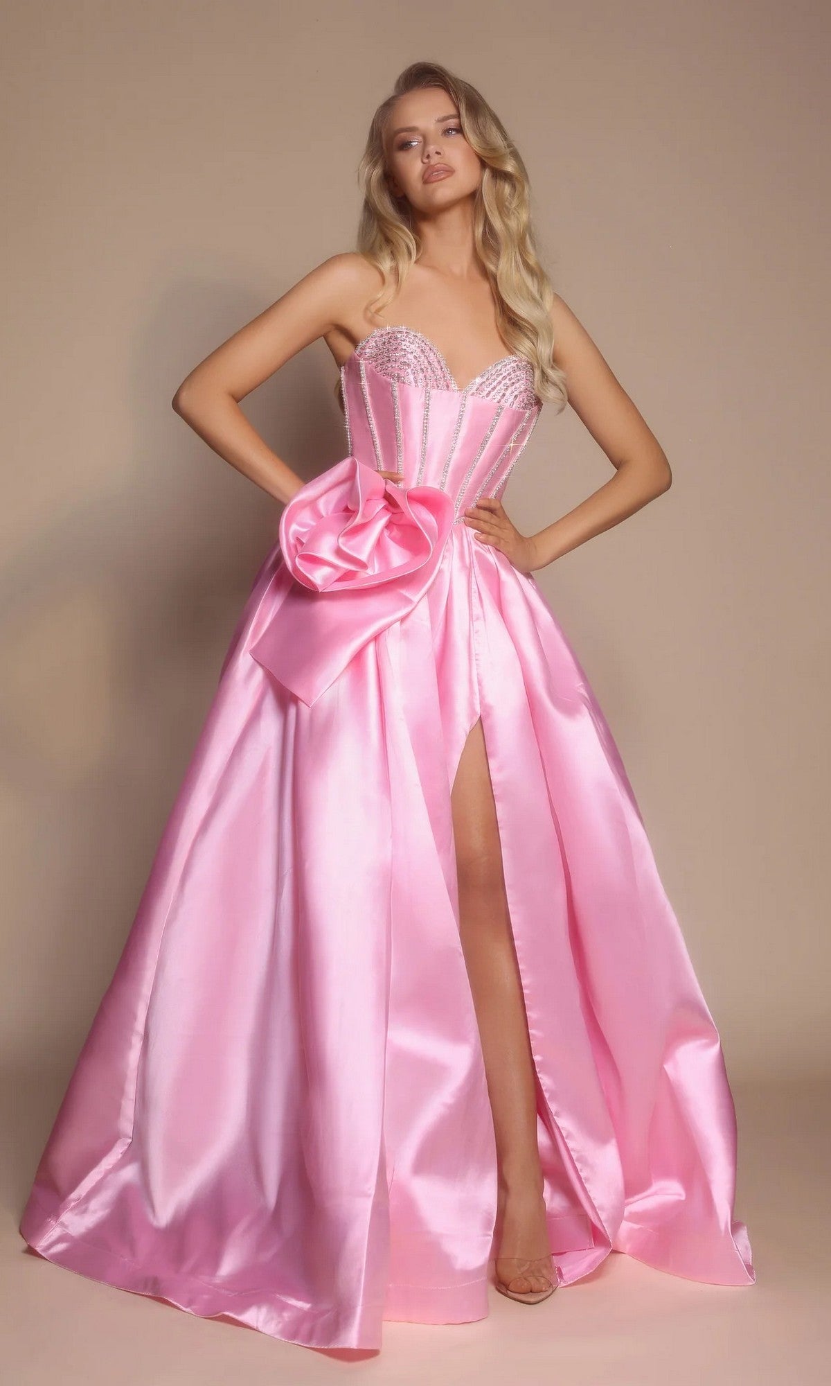 A woman in the Portia and Scarlett PS26079, a long pink formal dress with a strapless satin ball gown silhouette, beaded corset bodice, large bow detail, and thigh-high slit stands against a plain beige background.